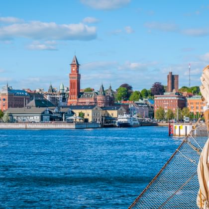 View of Helsingborg waterfront from a sailing ship. Red brick tower and historic buildings line the harbor under blue sky with white clouds.