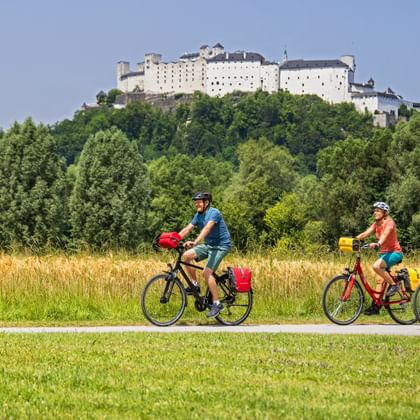 Three cyclists riding on a path through green meadows with Hohensalzburg Fortress on a wooded hill in the background under blue sky.