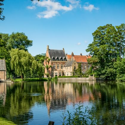 Minnewater Park in Bruges showing a tranquil lake reflecting historic buildings with red roofs and a church spire, surrounded by lush green trees.