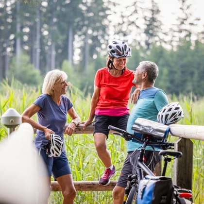 Three cyclists taking a break on a wooden fence in Chiemgau. Green field and forest in background. Two wear helmets, bikes visible.