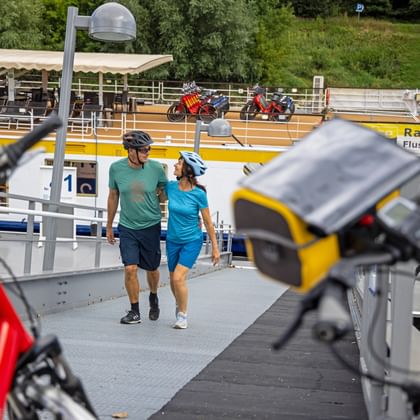 Two cyclists walking on boarding ramp of Danube river cruise ship in Melk. Bicycles visible in foreground, yellow ship railing with 'Rad & Schiff' text.