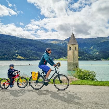 Erwachsener und Kind radeln am Uferweg des Reschensees mit dem berühmten versunkenen Kirchturm im türkisfarbenen Wasser, Berge im Hintergrund.