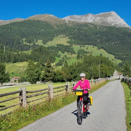 Radfahrerin auf asphaltiertem Radweg, umgeben von eingezäunten Wiesen, im Hintergrund Berge