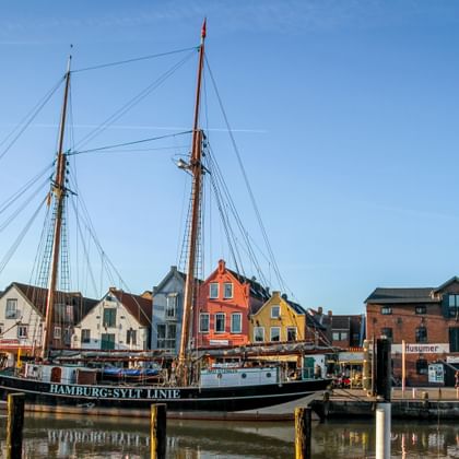 Traditionelles Zweimaster-Segelschiff im Hafen von Undine vor bunten historischen Gebäuden unter blauem Himmel.