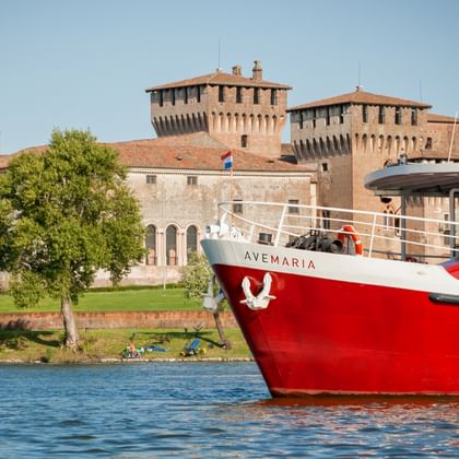Red and white passenger ship MS Ave Maria on water with historic fortress towers and green lawns of Mantua in the background under clear sky.