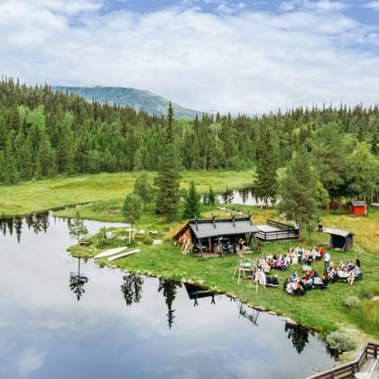 Luftaufnahme einer Versammlung am ruhigen See in Hemsedal Kommune. Menschen sitzen um eine Holzhütte, umgeben von grünem Wald und Bergen.
