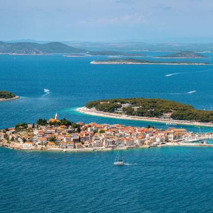 Aerial view of Primošten peninsula with terracotta roofs, church tower, and turquoise Adriatic Sea. Islands and mountains visible in background.