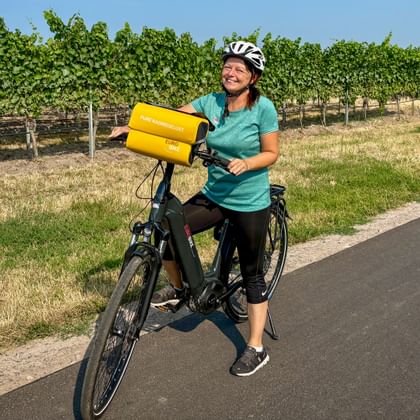 Radfahrerin mit Helm hält gelbe Fahrradtasche auf Radweg neben Weinbergreihen unter blauem Himmel in der Region Speyer-Heidelberg-Worms.
