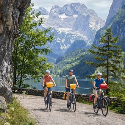 Drei Radfahrer fahren am Gosausee entlang mit dramatischen Dachstein-Gipfeln im Hintergrund. Felswand links, Alpensee und Wald sichtbar.