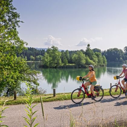 Drei Radfahrer fahren auf einem Schotterweg entlang eines ruhigen Sees in Kärnten. Grüne Bäume rahmen die Szene mit bewaldeten Hügeln im Hintergrund.