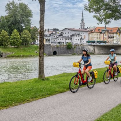 Three cyclists with yellow panniers riding along the Isar River in Bad Tölz. Historic buildings and a church spire visible across the river.