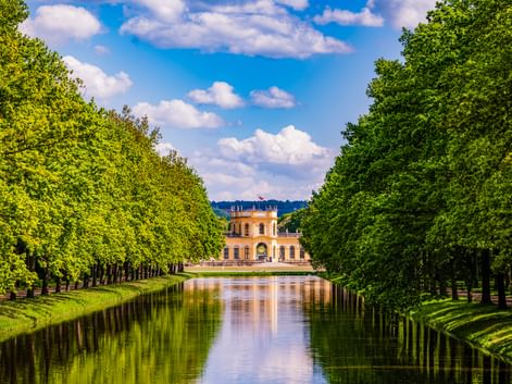 Gelbes barockes Orangerie-Gebäude in Kassel am Ende eines von Bäumen gesäumten Kanals. Grüne Bäume rahmen beide Seiten unter blauem Himmel.