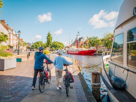 Zwei Radfahrer mit Rädern an einem Hafenkai, vor einem roten Feuerschiff im Wasser. Historische Gebäude und Kirchturm im Hintergrund.