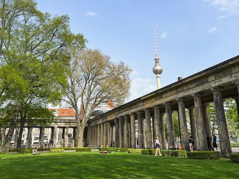Berlin TV Tower rising behind neoclassical colonnade with green lawn in foreground. Visitors walk through the columns on a sunny day.