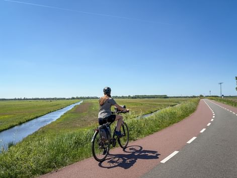 Radfahrerin mit Rucksack auf rotem Radweg entlang eines Kanals durch flache holländische Polderlandschaft unter blauem Himmel.