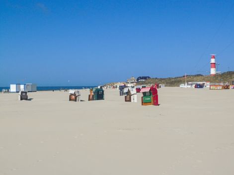 Sandstrand auf Borkum mit bunten Strandkörben, rot-weiß gestreiftem Leuchtturm im Hintergrund, blauer Himmel und Nordsee sichtbar.