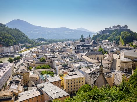 Panoramic view of Salzburg's old town with Hohensalzburg Fortress on the hill, baroque churches, and mountains in the background.