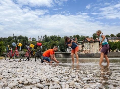 Three cyclists wading in the shallow Isar river with pebble beach. Four touring bikes with yellow bags parked on shore. Bad Tölz town visible in background.
