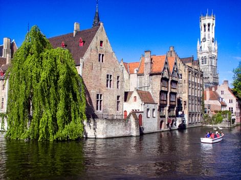 Mittelalterliche Gebäude entlang eines Kanals in Brügge mit dem Belfried-Turm. Eine Trauerweide hängt über das Wasser, ein Boot mit Touristen fährt vorbei.