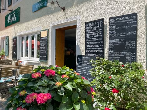 Outdoor café entrance with white textured walls, wooden door, and chalkboard menus. Colorful hydrangeas and flowers decorate the foreground.