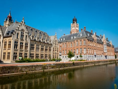 Historisches Rathaus Dendermonde mit verzierter roter Backsteinfassade und Turm neben Kanal unter blauem Himmel. Angrenzende Gebäude säumen das Ufer.