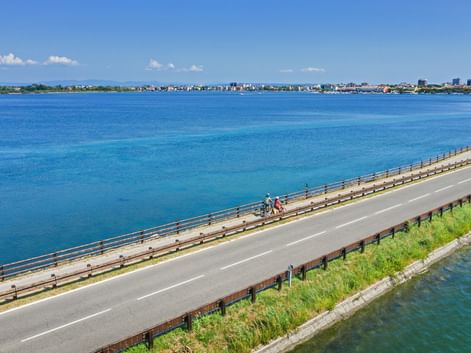 Aerial view of cyclists on a bridge crossing a turquoise lagoon near Grado, Friuli. The coastal town is visible in the distance under blue sky.