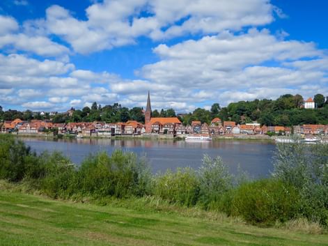 Blick auf die Stadt Lauenburg mit rotgedeckten Gebäuden und Kirchturm entlang der Elbe, eingerahmt von grüner Vegetation unter blauem Wolkenhimmel.