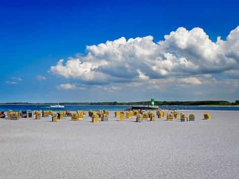 Sandstrand in Travemünde mit Reihen gelber Strandkörbe. Grüner Leuchtturm in der Ferne unter blauem Himmel mit weißen Wolken.