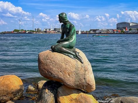 Bronze statue of The Little Mermaid sitting on a rock in Copenhagen harbor, with blue water and city skyline in the background.
