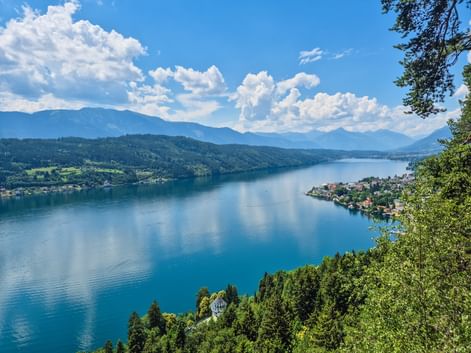 Panoramablick auf den Millstätter See von der Luschan Höhe Plattform. Blauer See umgeben von grünen Wäldern, kleine Dörfer am Ufer, Berge im Hintergrund.
