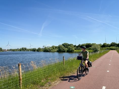 Radfahrerin auf rot gepflastertem Radweg entlang blauem Wasserweg mit grünem Gras, Bäumen und Windrädern unter klarem Himmel.