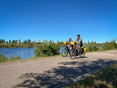 Zwei Radfahrer mit gelben Taschen auf asphaltiertem Weg neben Fluss mit grüner Vegetation und Zypressen unter blauem Himmel.