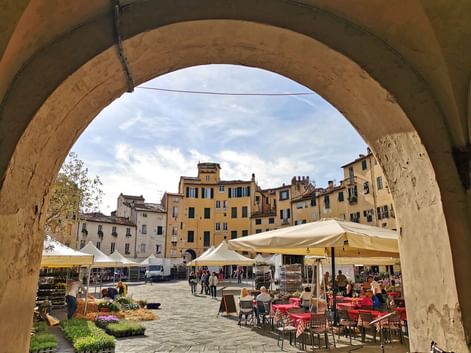View through stone archway to historic square in Lucca with colorful buildings, outdoor cafés, market stalls, and people under cloudy sky.
