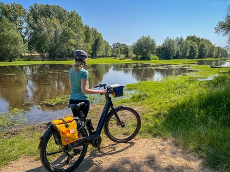 Female cyclist with helmet standing by e-bike with yellow panniers beside calm river. Green meadows and trees line the waterway under blue sky.