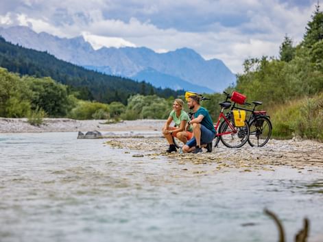 Zwei Radfahrer sitzen an einem Kiesstrand an der Isar mit beladenen Tourenrädern. Berge und Wälder im Hintergrund sichtbar.