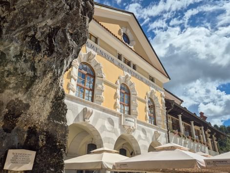 Gelbes Barockgebäude mit weißen Steinbögen und lateinischer Inschrift in Postojna. Eine große Felsformation ist links sichtbar.