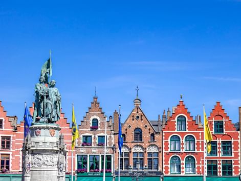 Historischer Grote Markt in Brügge mit bunten Giebelhäusern und einem Bronzedenkmal unter blauem Himmel. Fahnen schmücken die Gebäude.