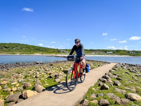 Cyclist on red bike riding along narrow coastal path surrounded by rocks and green vegetation, with blue water and coastline in background.