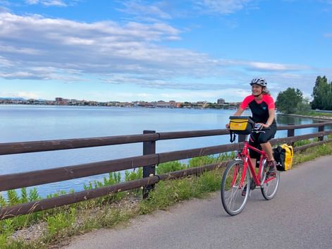 Radfahrer mit rotem Fahrrad und gelben Taschen auf Uferweg in Friaul, mit Holzgeländer, ruhiger Lagune und Küstenstadt im Hintergrund.