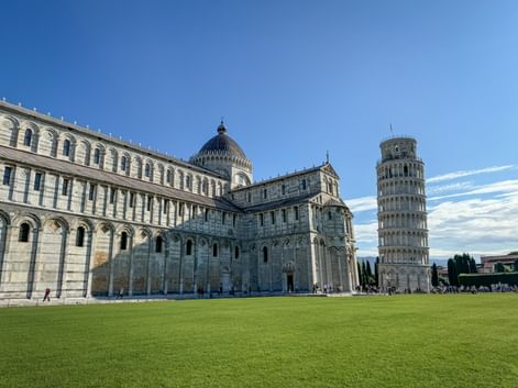Pisa Cathedral (Santa Maria Assunta) with its striped marble facade and the famous Leaning Tower on green lawn under blue sky.