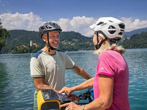 Two cyclists in helmets conversing by Lake Bled with forested mountains and church tower in background under blue sky with clouds.