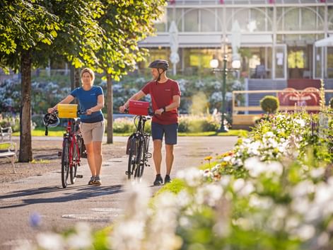 Zwei Radfahrer schieben ihre Räder durch einen sonnigen Park in Bad Ischl. Weiße Blumen im Vordergrund, Kongresshaus im Hintergrund sichtbar.