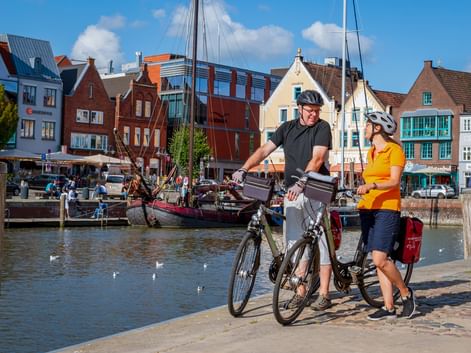 Zwei Radfahrer mit Rädern am Husumer Hafen. Bunte historische Häuser und Boote säumen die Uferpromenade unter blauem Himmel.