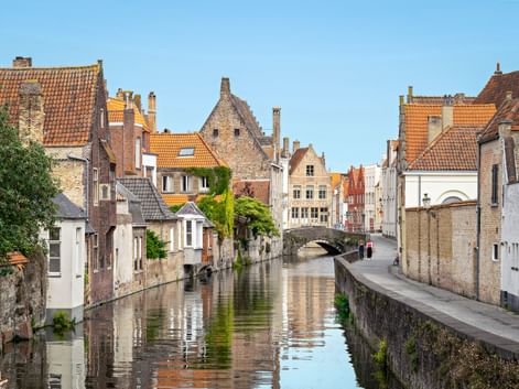 Picturesque canal in Bruges with medieval brick houses, orange-tiled roofs, and a stone bridge. Trees line the waterway under a clear blue sky.