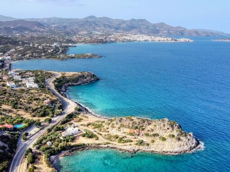 Luftaufnahme der Küste Kretas mit kurvenreicher Straße entlang türkiser Buchten, felsigen Halbinseln und Bergen im Hintergrund.