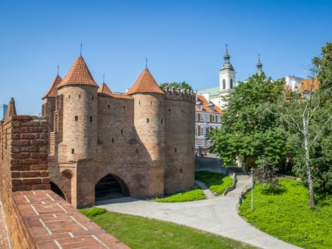 Warschauer Barbakan-Festung mit roten Ziegeltürmen und Kegeldächern. Grüne Rasenhänge und eine Kirche mit weißen Türmen im Hintergrund.