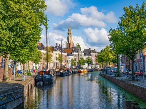Gracht in Groningen mit historischen Booten, bunten Häusern und hohem Kirchturm unter blauem Himmel mit weißen Wolken.