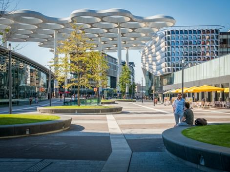 Utrechter Bahnhofsvorplatz mit markanter weißer Überdachung und geschwungenem modernen Gebäude. Menschen entspannen auf Bänken nahe Bäumen.