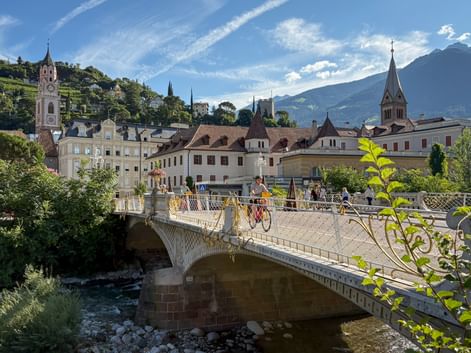 Radfahrer überqueren eine verzierte Steinbrücke über einen Fluss in Meran, mit historischen Gebäuden, Kirchtürmen und Bergen im Hintergrund.