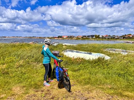 Cyclist with bike on grassy coastal area overlooking water and village houses in Träslövsläge, Sweden. Blue sky with white clouds above.